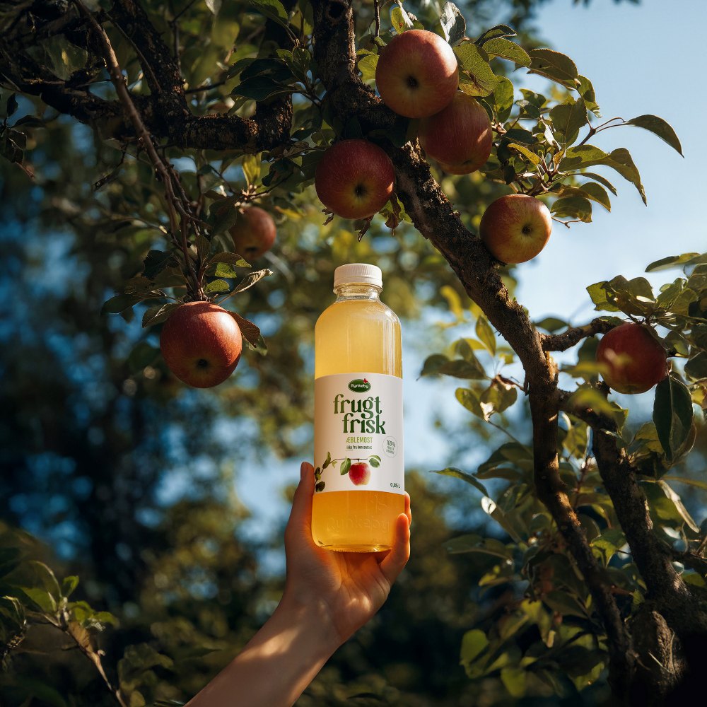 A hand holds a bottle of fruit juice labeled "frugt frisk" under an apple tree with ripe apples against a clear blue sky.