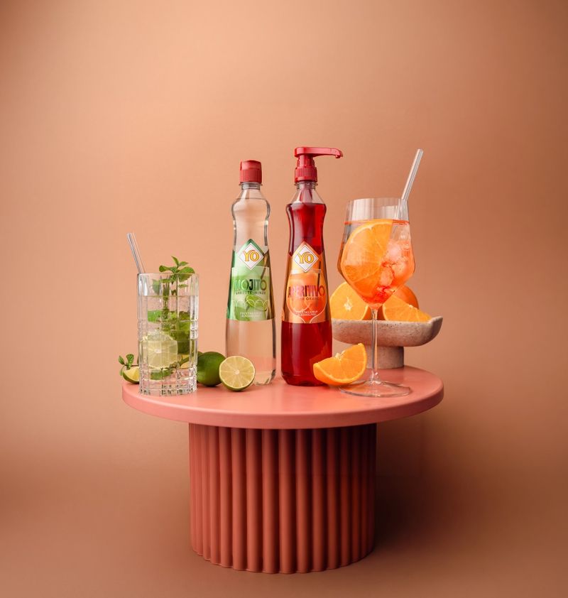 Two bottles of Cocktailsirup YO Fruchtsirup, garnished cocktails, mint, lime, orange slices on a terracotta pedestal against a peach background.