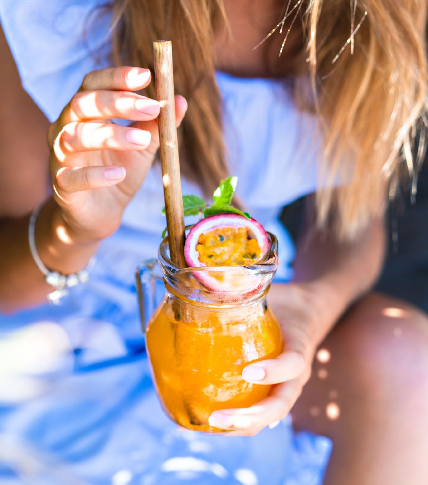 A person in a light blue dress holds a glass jar of orange drink with a bamboo straw, garnished with passion fruit and mint.