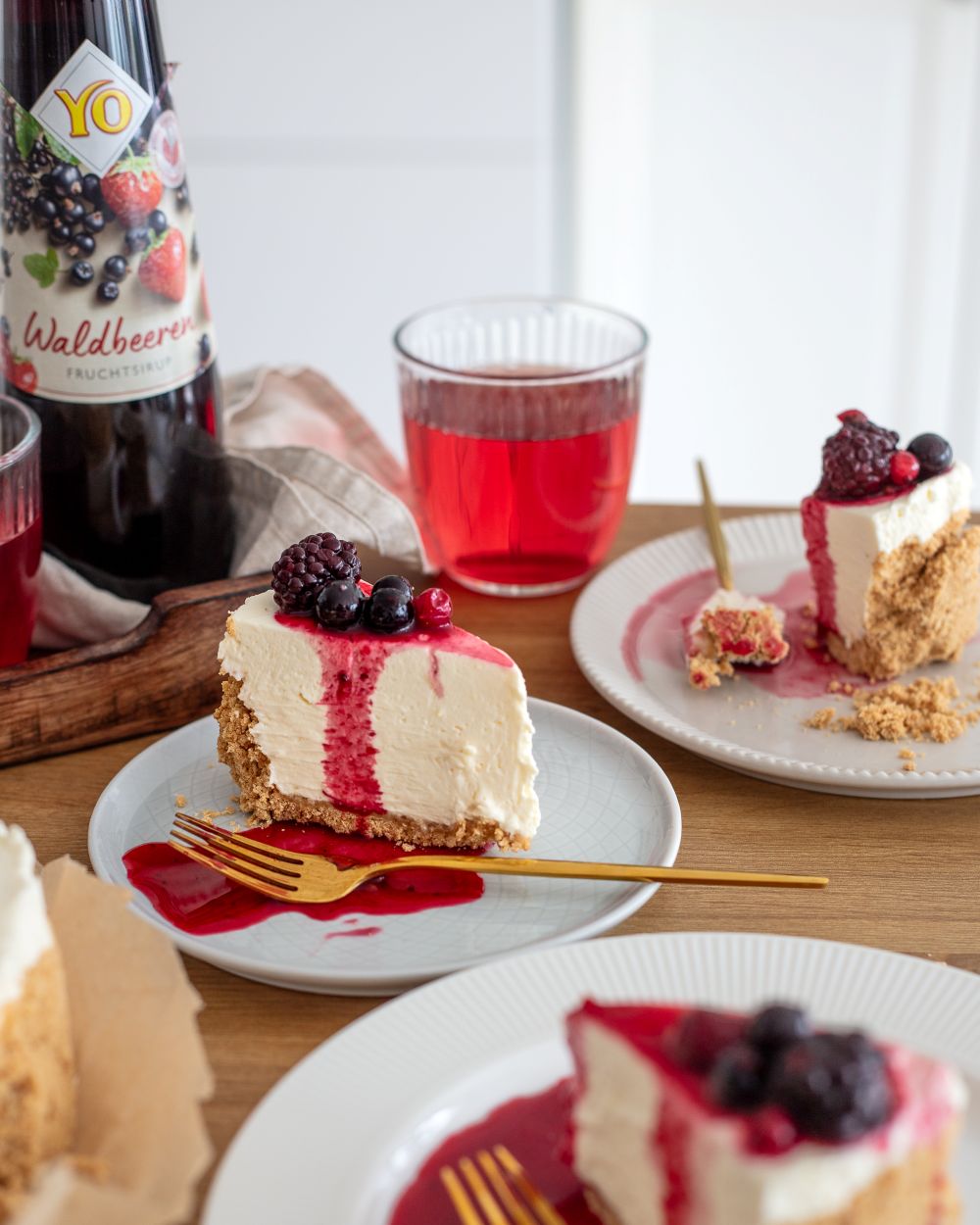 Slice of cheesecake with berry topping and sauce on a plate, beside a glass of red drink and a bottle, on a wooden table.