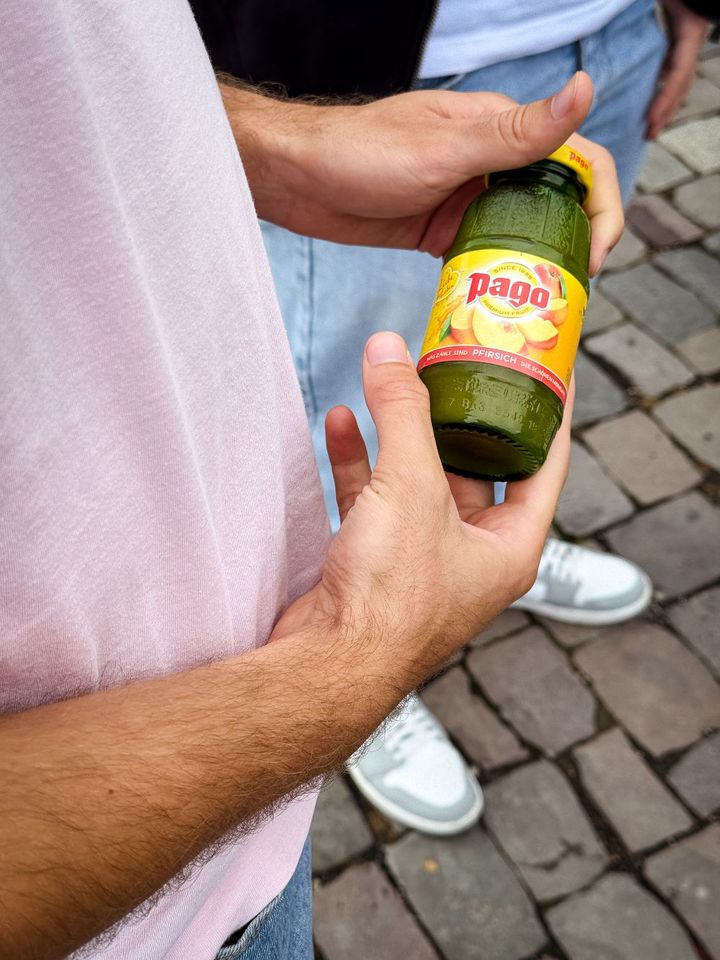 Person holding a bottle of orange Pago juice on a cobblestone street, wearing a pink shirt and blue jeans.