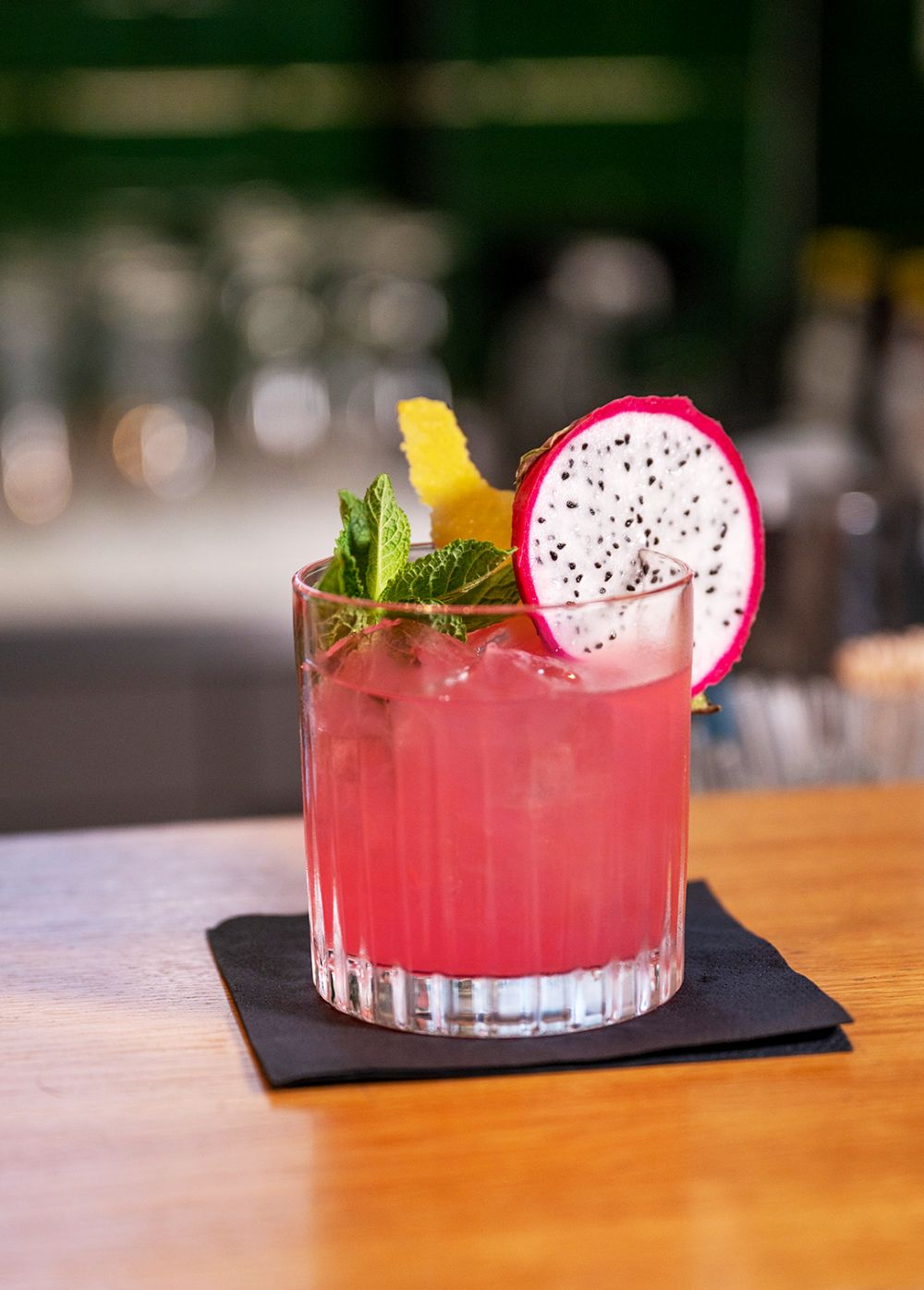 A colorful cocktail in a glass with ice, garnished with a slice of dragon fruit, mint leaves, and a lemon twist, on a black napkin.
