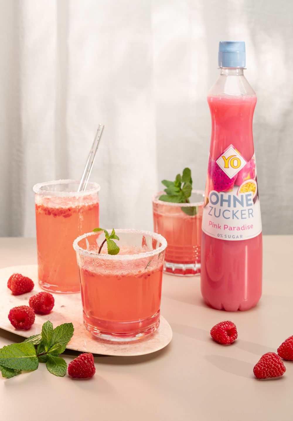 Three glasses of pink raspberry drink with mint garnish sit beside a YO bottle labeled "Ohne Zucker," surrounded by fresh raspberries and mint.