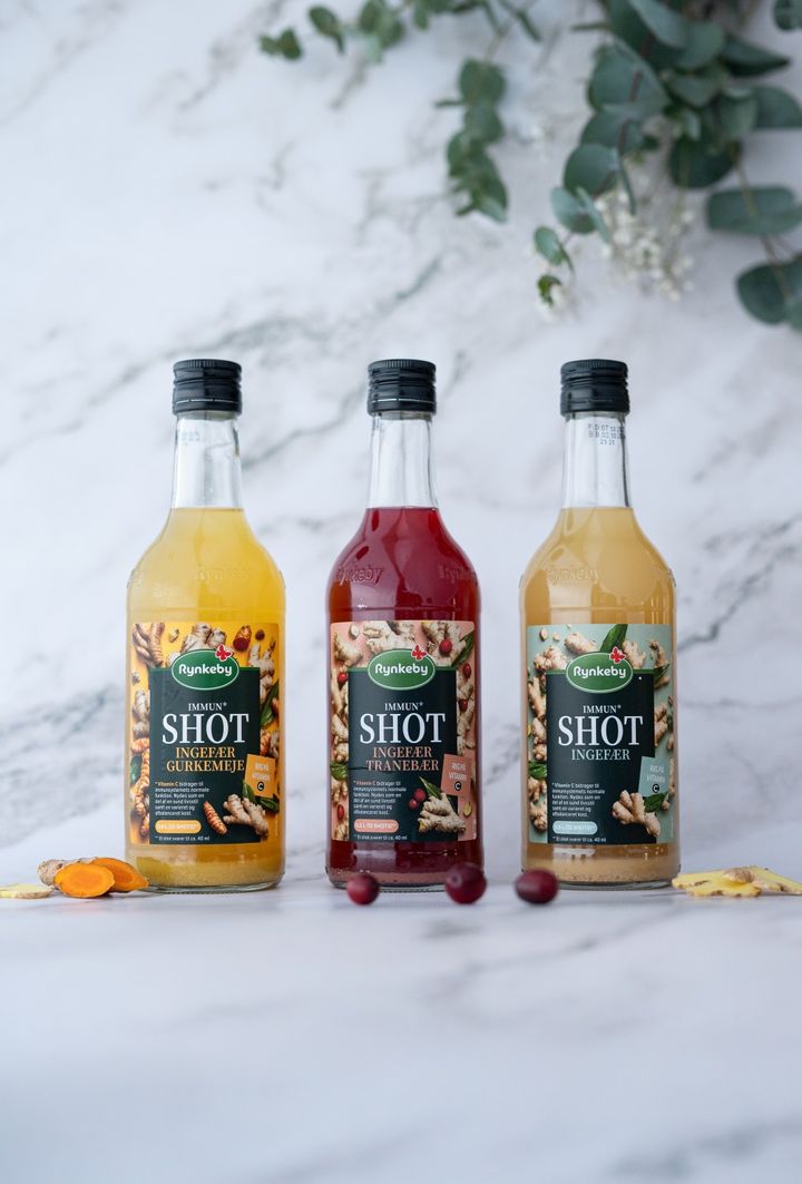 Three bottles of Rynkeby Immun+ ginger shots on a marble surface, surrounded by cranberries and turmeric slices.