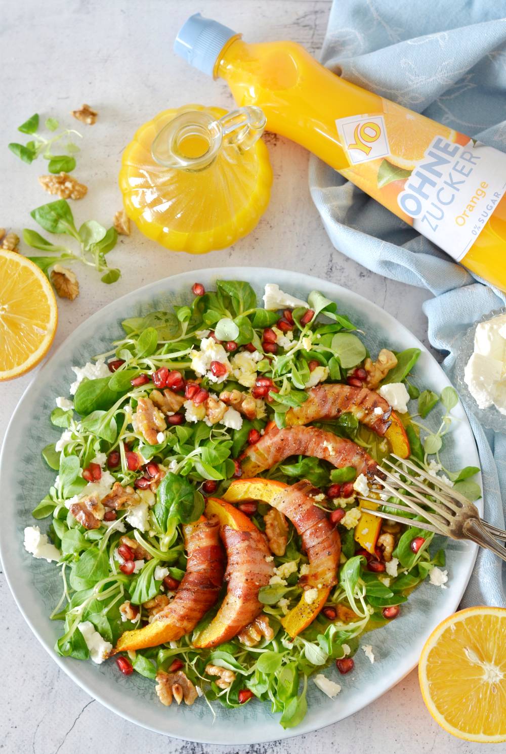 A vibrant salad with greens, grilled peaches, walnuts, pomegranate seeds, and cheese crumbles, served with a yellow sauce bottle on the side.