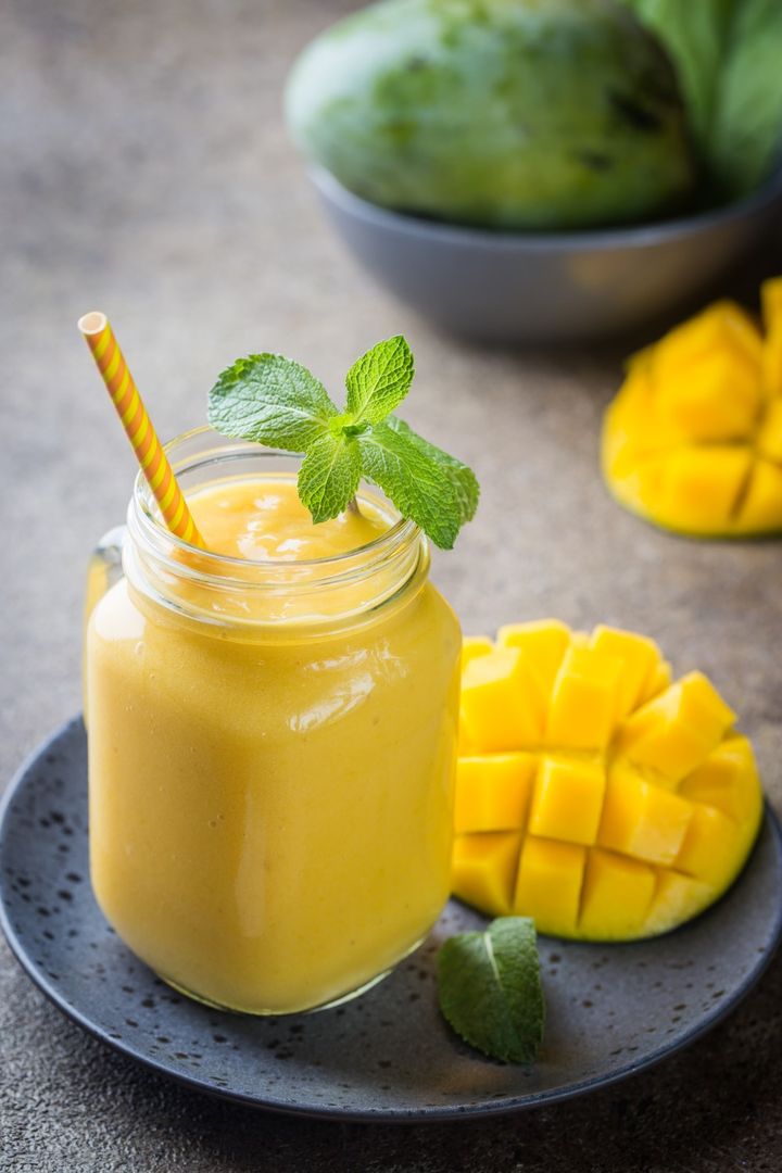 A mango smoothie in a jar with a striped straw and mint garnish, on a plate with diced mango pieces, and a whole mango in the background.