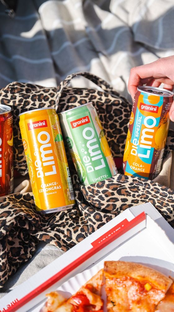 Cans of Granini Die Limo drinks on a leopard print cloth beside a pizza box, with a hand reaching for one can.