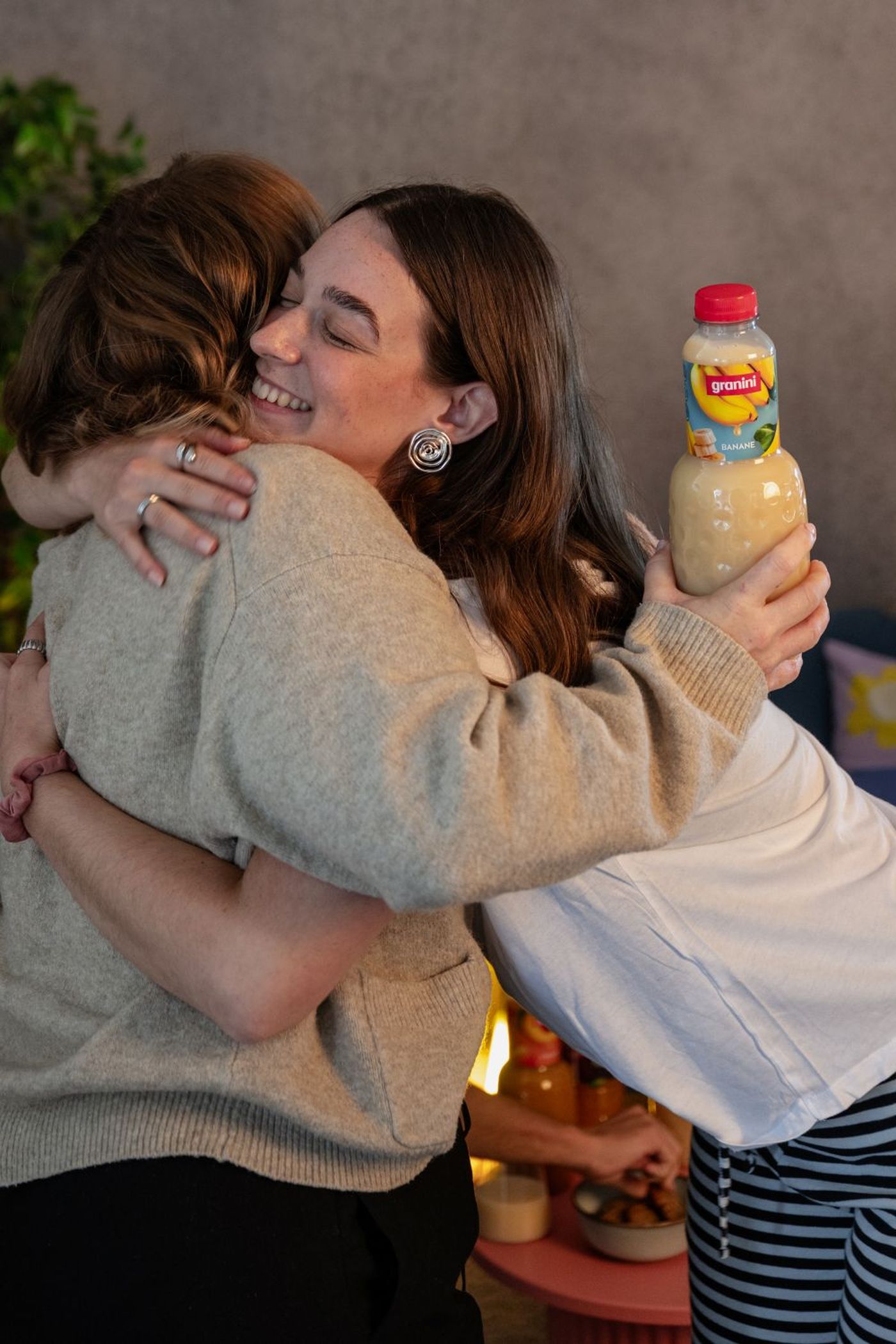 Two people hugging warmly, one holding a bottle of banana juice labeled "granini," with snacks in the background.