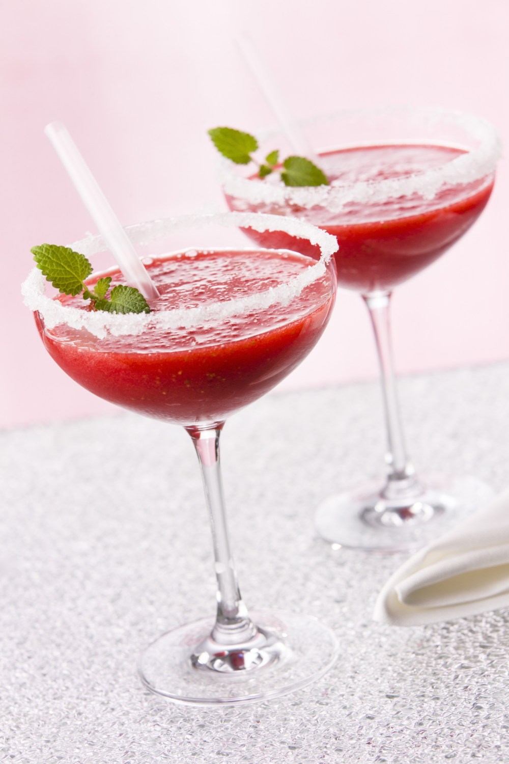Two strawberry margaritas in sugar-rimmed glasses with mint leaves and straws, set against a pink background.