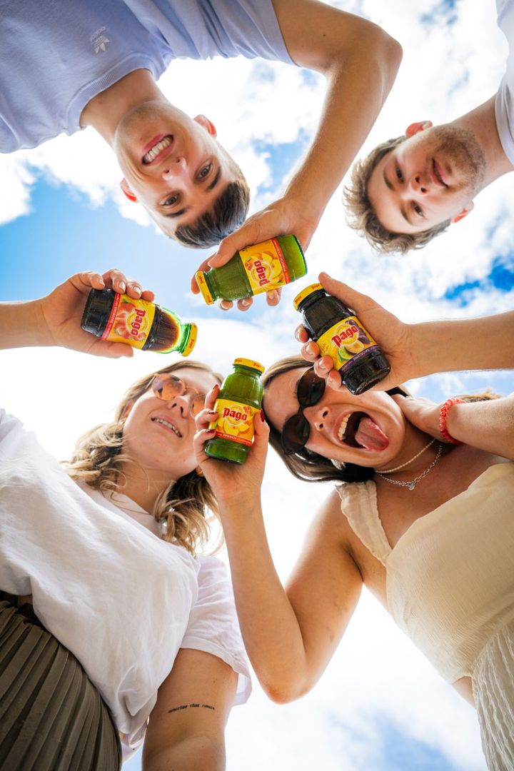 Four people smiling down at the camera, holding up bottles of Pago juice against a bright blue sky.