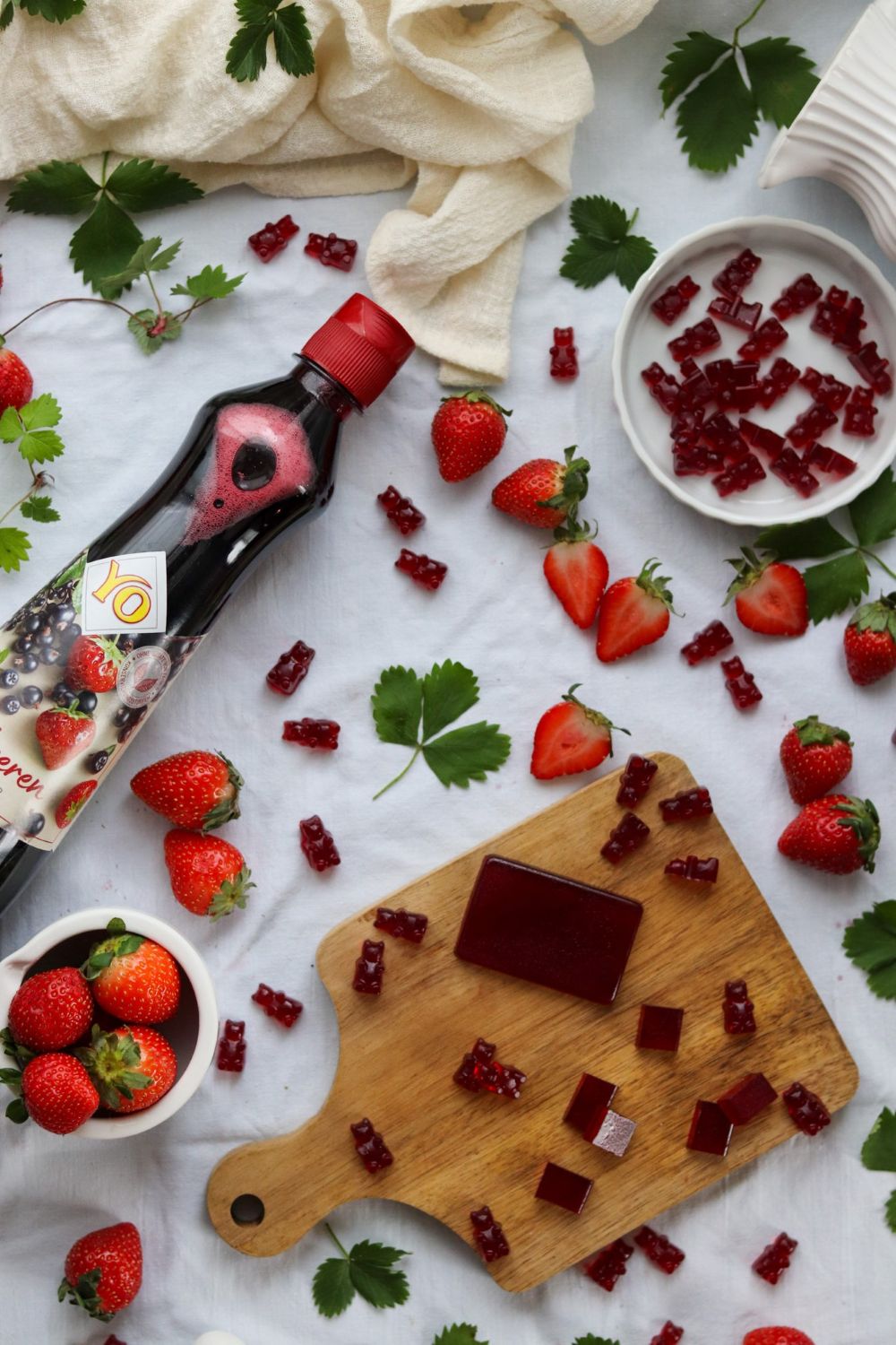 A bottle of fruit juice lies on a white cloth, surrounded by strawberries, gummy candies, and fresh parsley on a wooden cutting board.