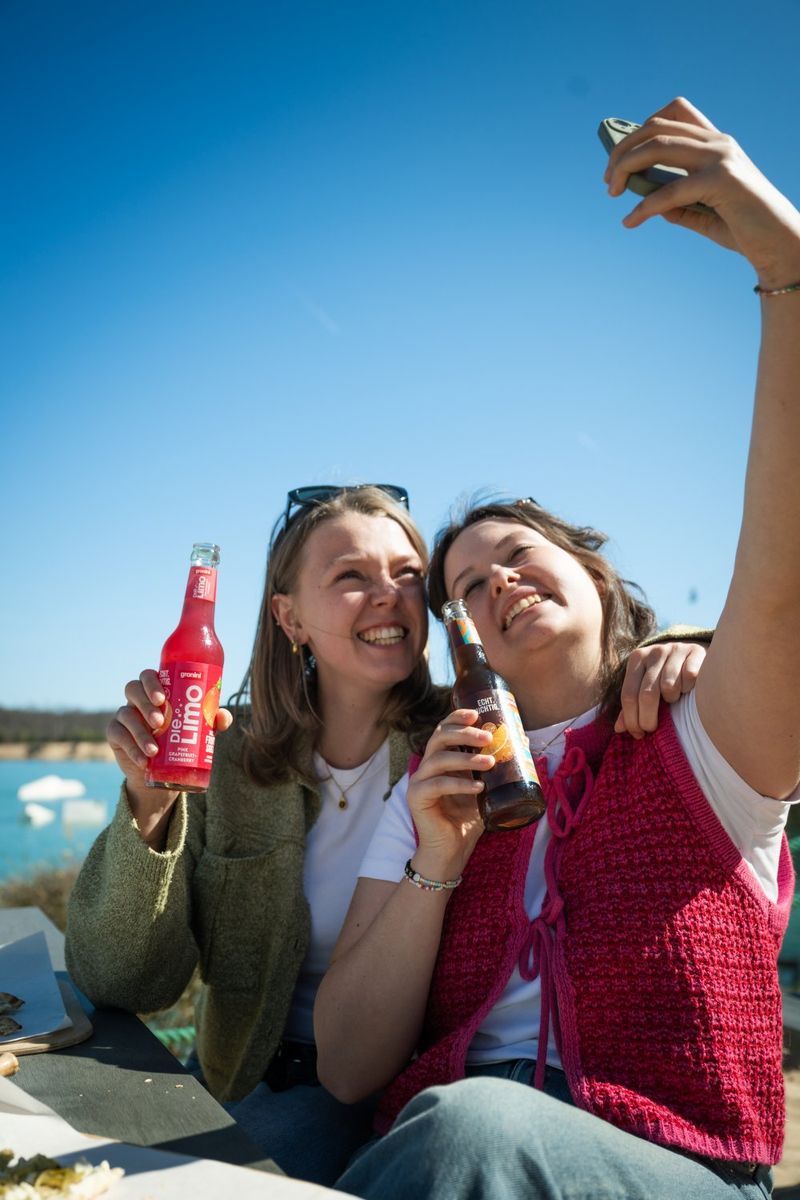Two friends taking a selfie, smiling and holding drinks, outdoor picnic setting with a clear blue sky in the background.