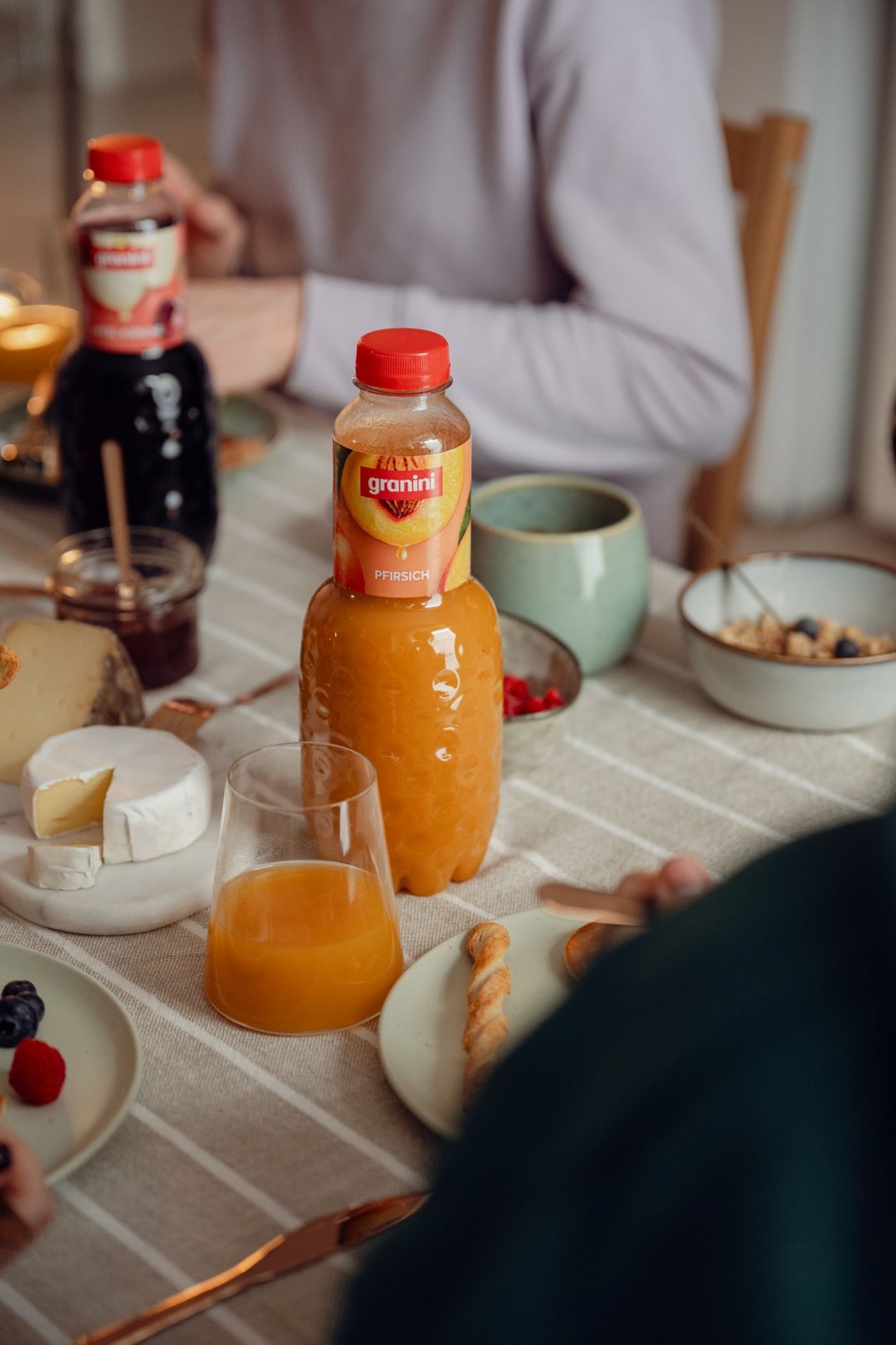 Breakfast table with a bottle of peach juice, orange juice glass, cheese, breadstick, berries, and bowls on a striped tablecloth.