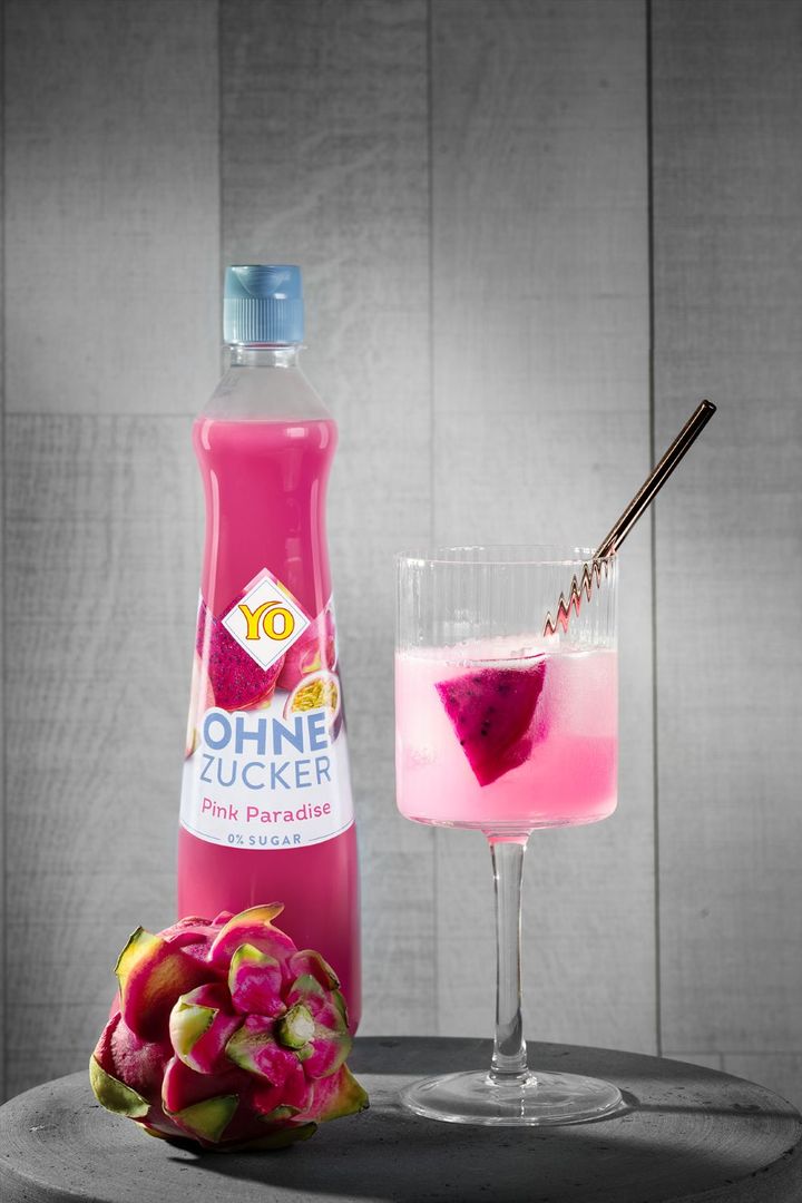 A bottle of "YO Ohne Zucker Pink Paradise" drink next to a glass of pink liquid with a dragon fruit slice, against a wooden background.