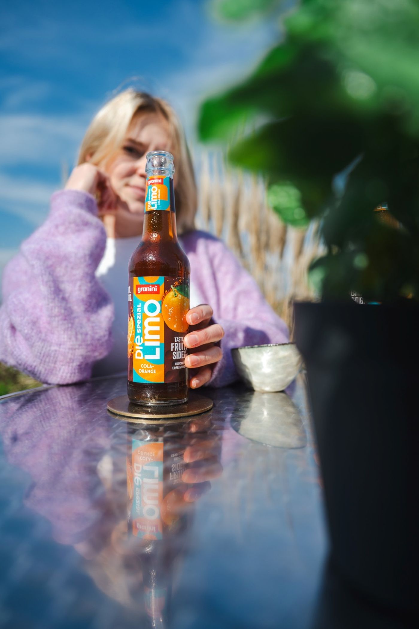 Person in a purple sweater holding a bottle of Granini Limo on a reflective table outdoors, with blue sky and blurred greenery in the background.