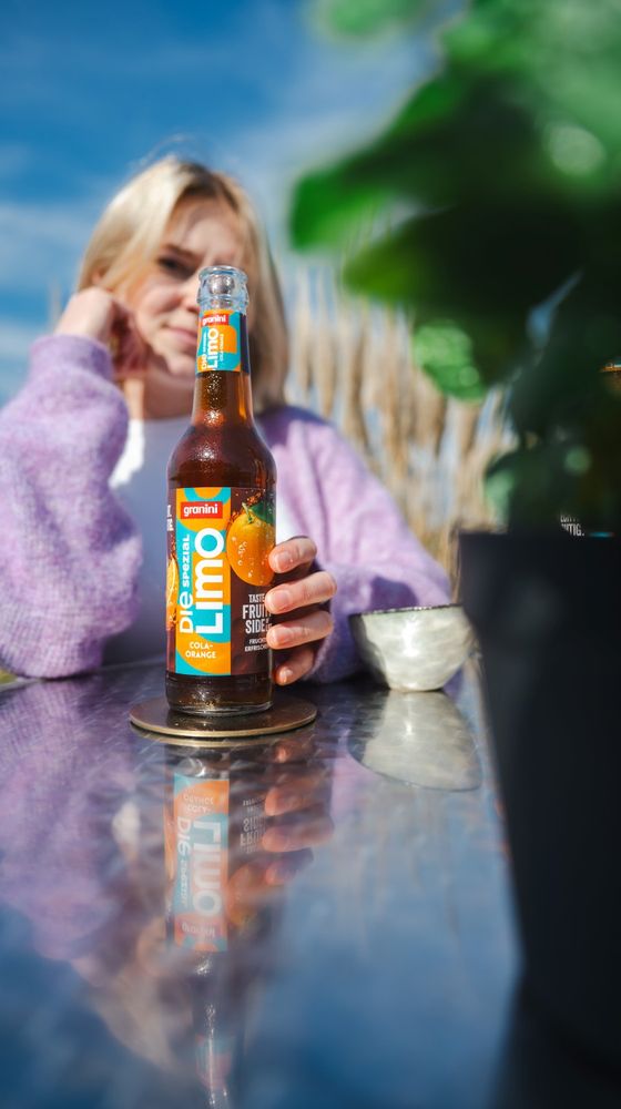 Person in a purple sweater holding a bottle of Granini Limo on a reflective table outdoors, with blue sky and blurred greenery in the background.