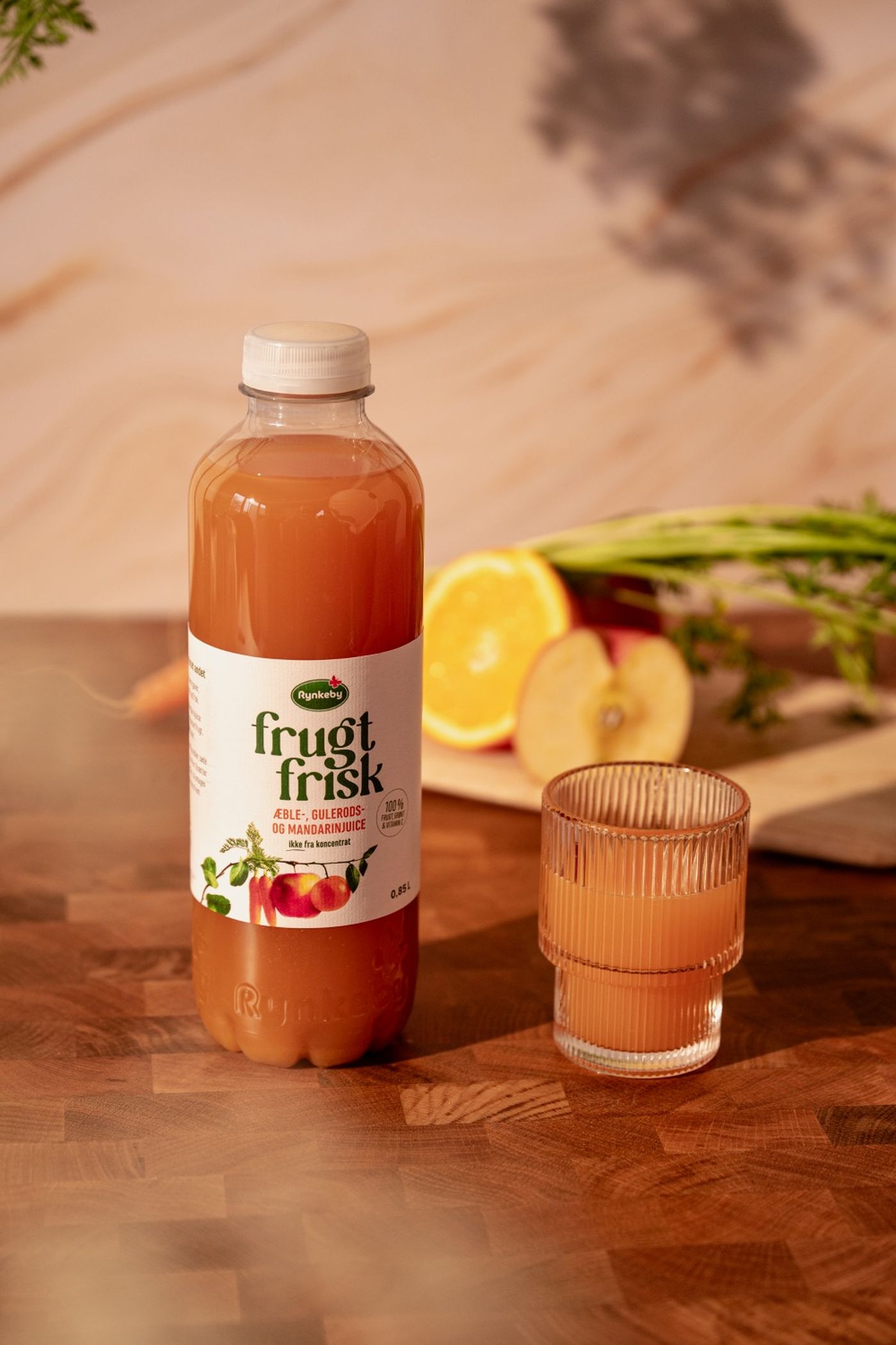 A bottle of Rynkeby fruit juice and a glass beside it, with sliced oranges and apples in the background on a wooden table.