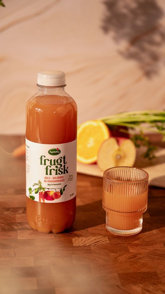 A bottle of Rynkeby fruit juice and a glass beside it, with sliced oranges and apples in the background on a wooden table.
