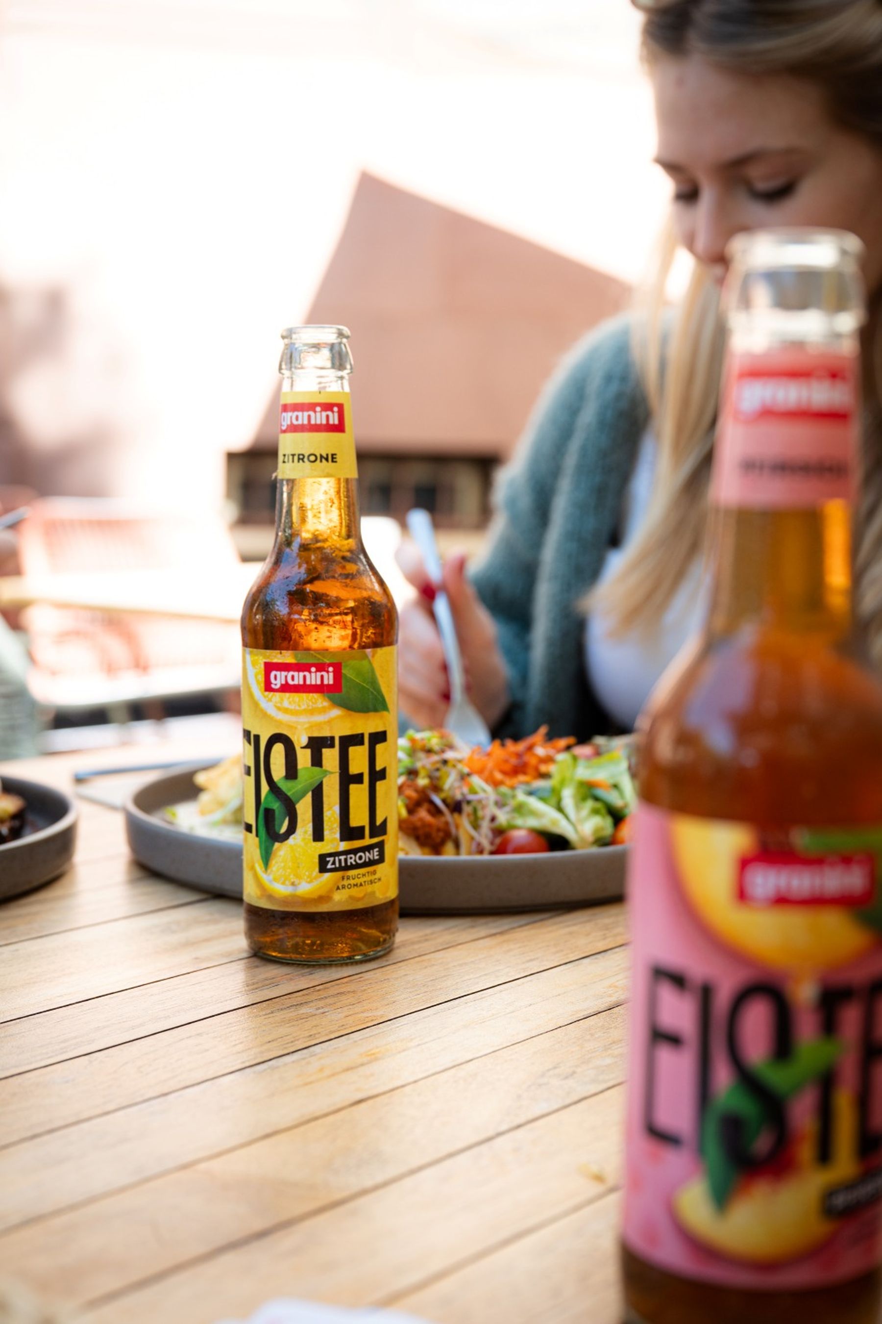 Bottle of iced tea on a wooden table with a woman eating a salad in the background.