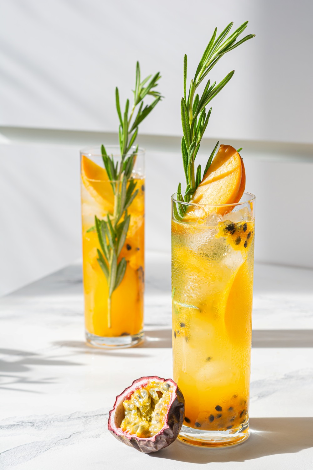 Two tall glasses of orange passion fruit cocktails with rosemary garnish on a sunny surface, accompanied by a halved passion fruit.