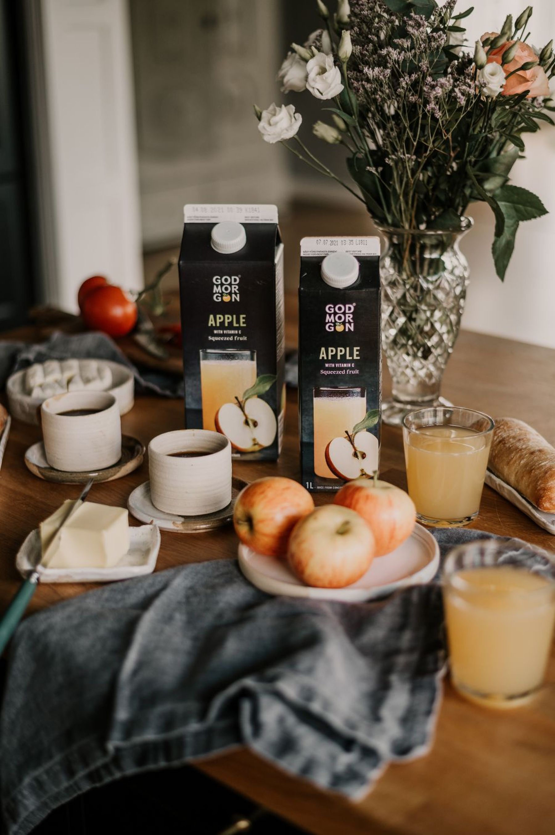 A breakfast table with apple juice cartons, apples, juice in glasses, and a vase of flowers on a wooden surface.