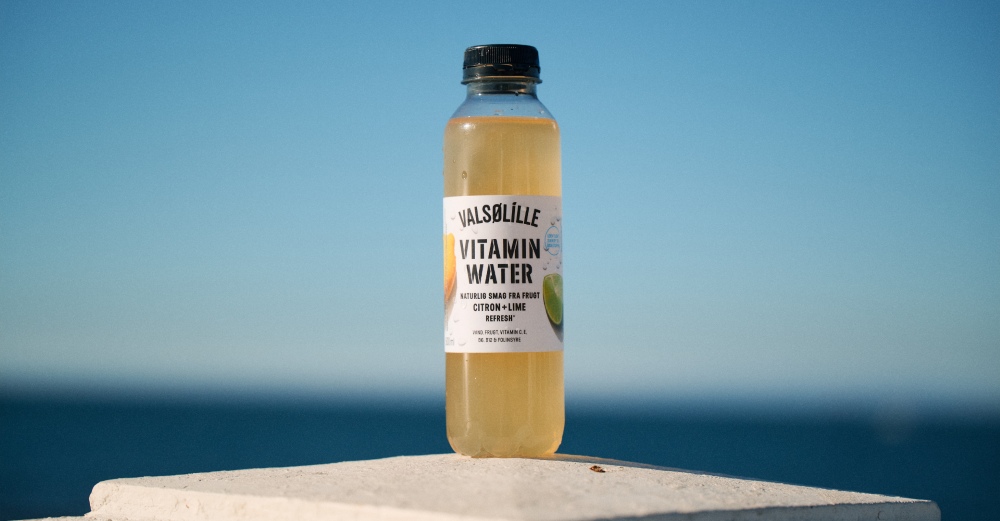 A bottle of Valsølille vitamin water with lemon-lime flavor on a ledge against a clear blue sky and sea backdrop.