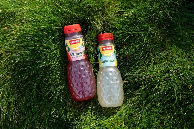 Two bottles of granini citronnade on green grass, one red and one clear, with textured surfaces and red caps.