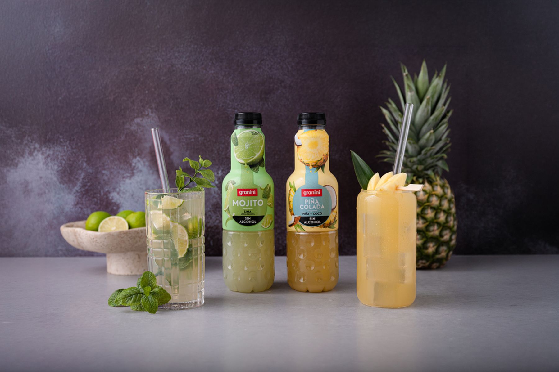 Two bottles of non-alcoholic mojito and piña colada drinks with garnished glasses, a bowl of limes, and a pineapple on a gray surface.
