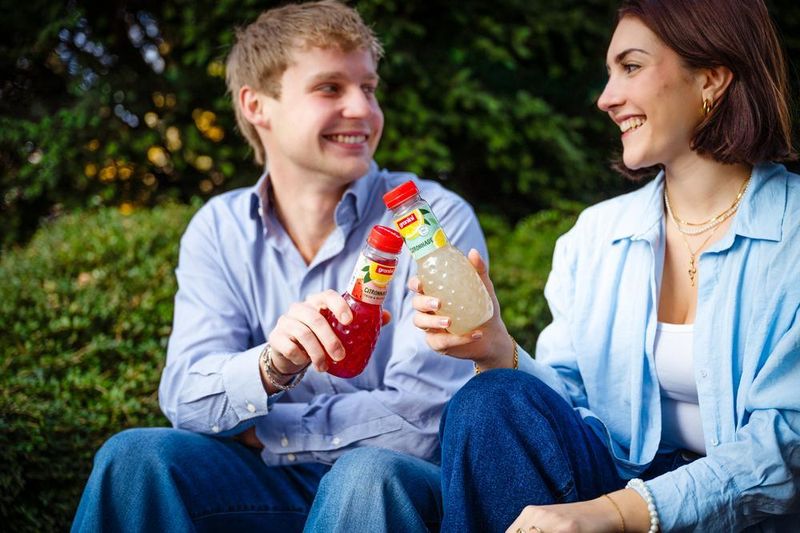 A man and woman smiling at each other while sitting outdoors, each holding a bottle of flavored drink.