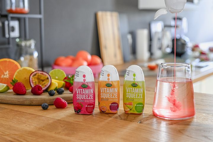 Vitamin Squeeze bottles on a wooden table with fresh fruit. Red liquid pours into a glass of water, creating a vibrant swirl effect.