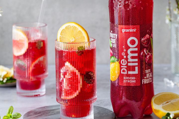 Tall glass of red cherry-lemon soda with lemon slice, next to a bottle labeled "Die Limo" on a gray surface.