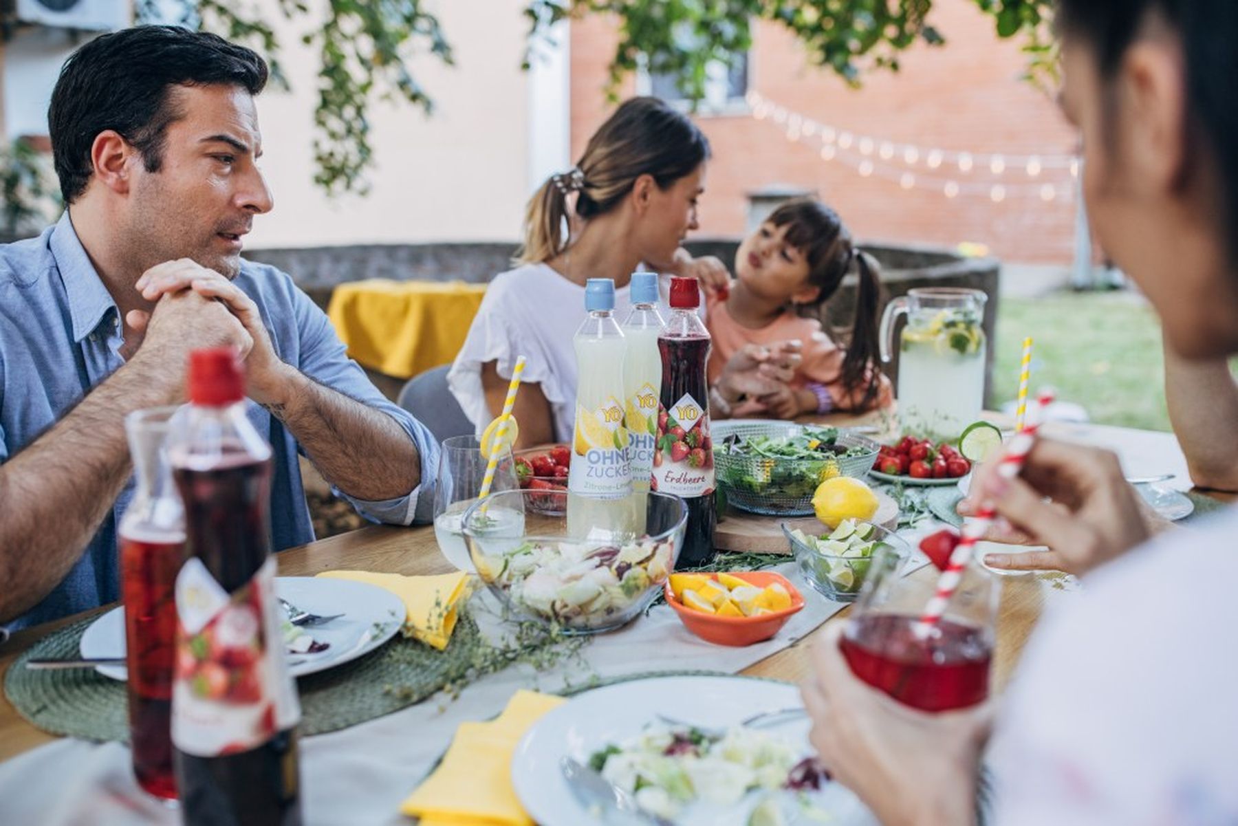 A family enjoys a summer meal outdoors with drinks, fresh fruit, and salads on a wooden table, surrounded by greenery and string lights.