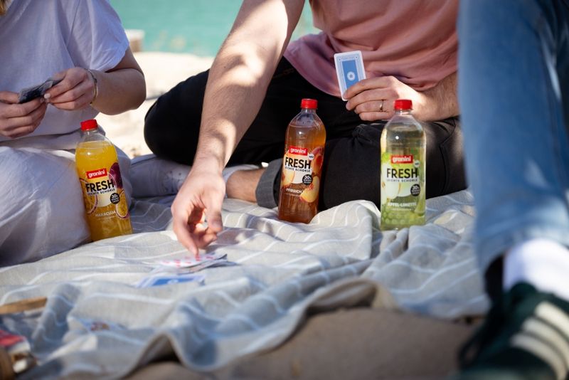 People playing cards on a picnic blanket with three bottles of granini Fresh drinks placed nearby.