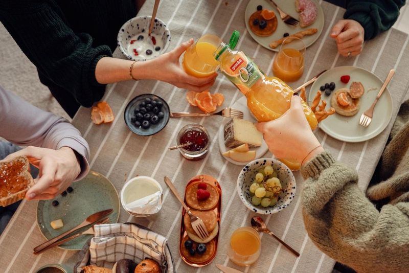 A table with brunch items: granini, fruits, cheese, jam, pastries, and people pouring juice. Plates and hands