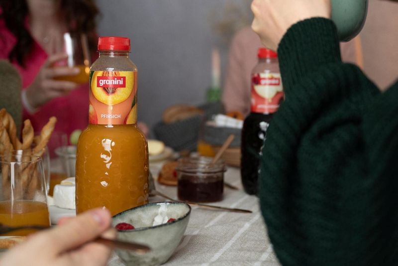 Close-up of a breakfast table with a bottle of peach juice, breadsticks, and jam. People are holding cups and glasses, with a casual setting.