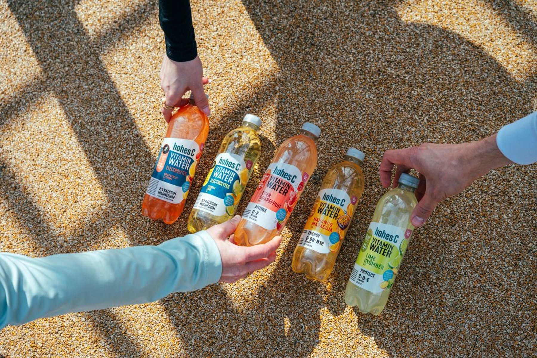 Hands reach for various Hohes C Vitamin Water bottles in assorted flavors, displayed on a pebbled surface under sunlight.