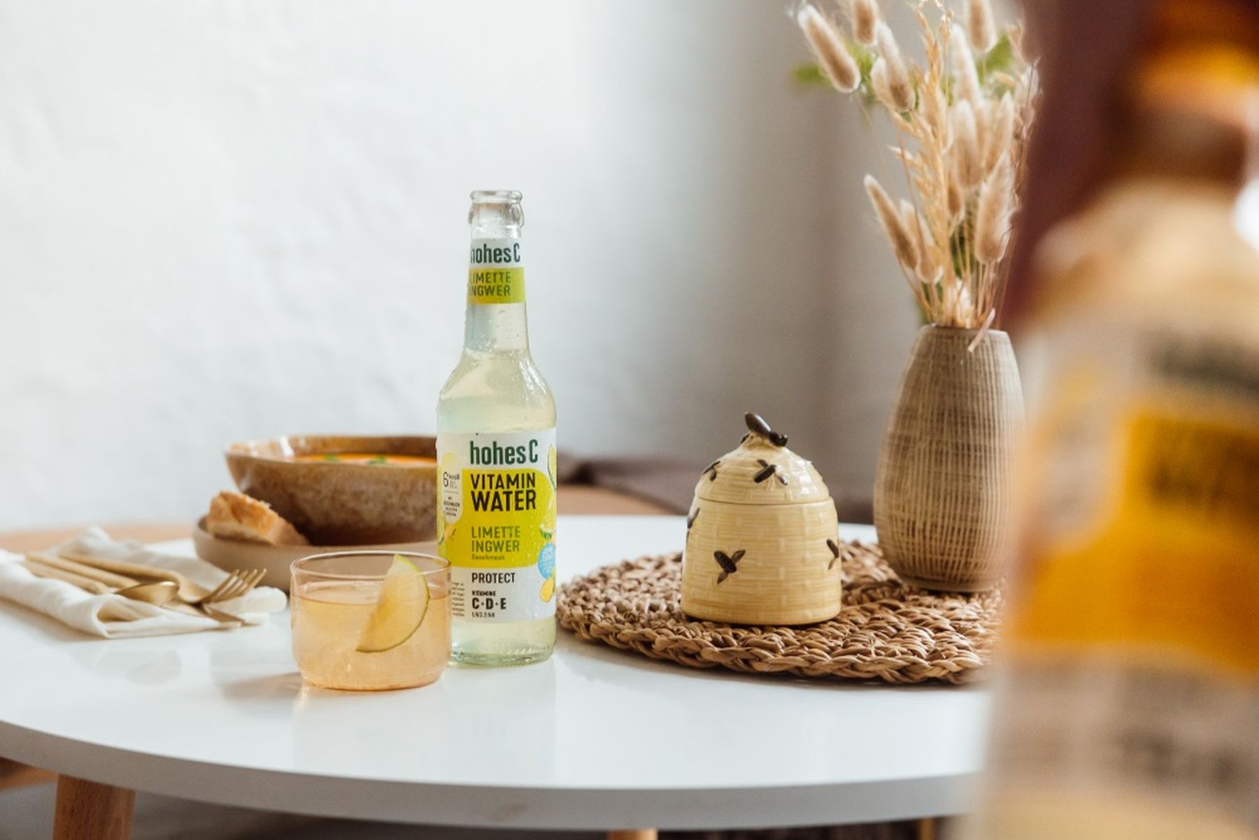 A bottle of hohes C Vitamin Water and a drink with lemon on a white table, next to a vase with dried flowers and a decorative ceramic jar.