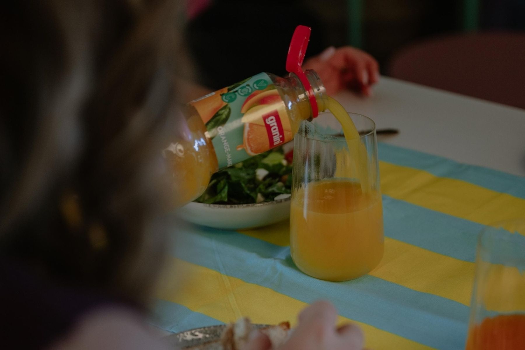 A person pours orange juice from a bottle into a glass over a blue and yellow tablecloth. A bowl of salad is on the table.