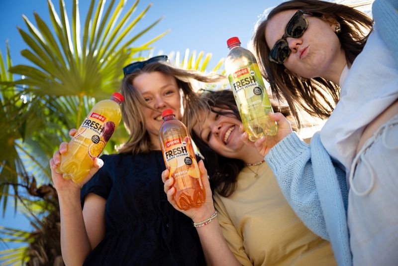 Three people smiling and holding fruit juice bottles, standing under palm trees with a clear blue sky in the background.