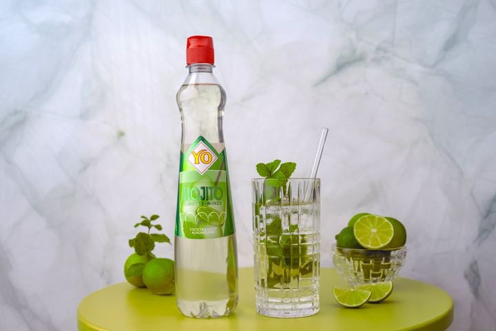 Bottle of YO mojito syrup beside a glass filled with the drink, mint, and ice on a green table with limes in a bowl.