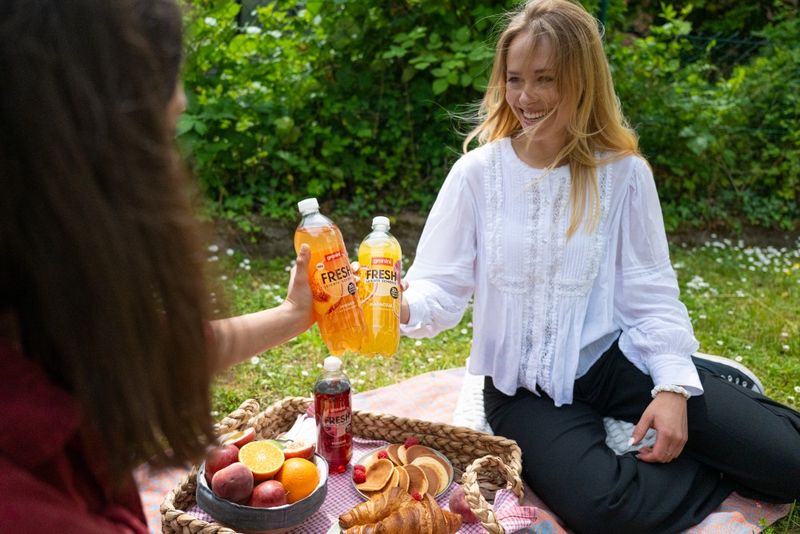 Two people at a picnic toast with granini FRESH. One holds orange juice, the other grapefruit. A basket with fruit and croissants sits nearby.