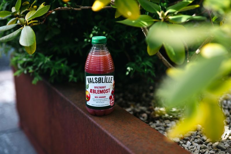 A bottle of "Valsølille Unfiltered Æblemost" with raspberries, placed on a brown ledge next to green foliage.