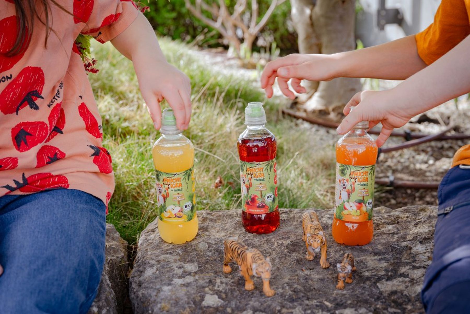 Two children sitting on rocks, each holding a colorful Fruchttiger. Toy tigers are placed in front of them.