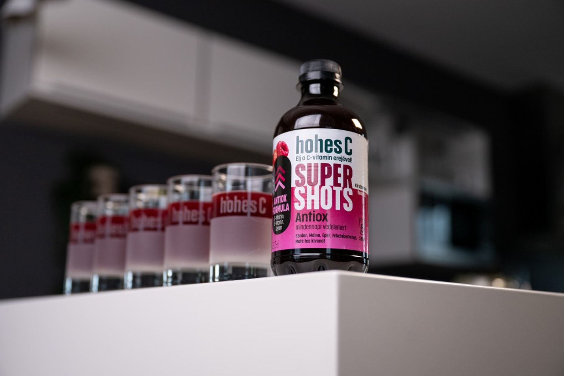 Bottle of Hohes C Super Shots Antiox on a white surface, with a row of branded glasses filled with pink liquid in the background.