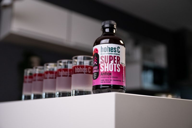 Bottle of Hohes C Super Shots Antiox on a white surface, with a row of branded glasses filled with pink liquid in the background.