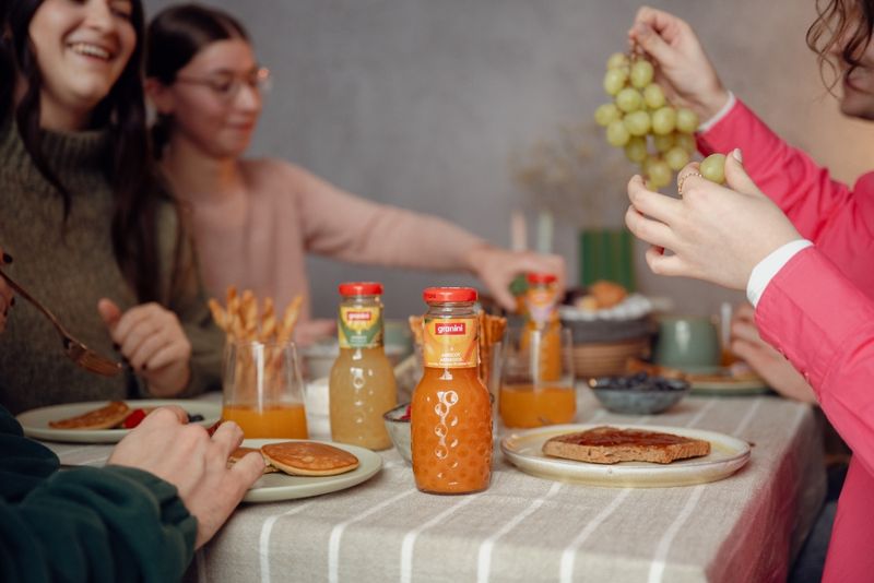 People enjoying a meal at a table with pancakes, juice, and grapes. Bottles of apricot juice are prominently displayed on the table.