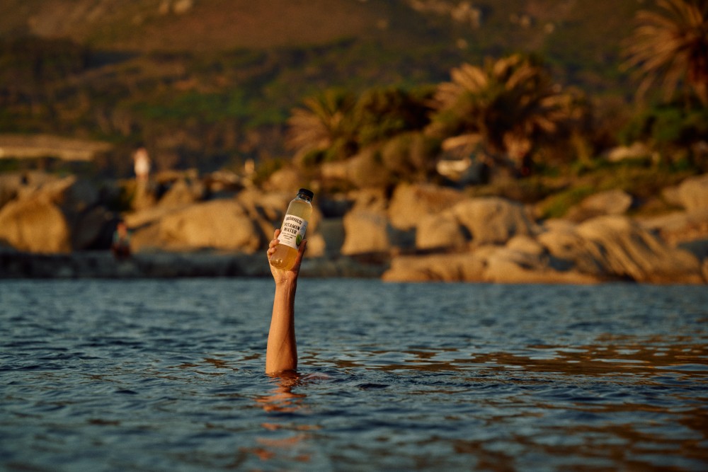A person in water holds up a bottle, surrounded by large rocks and palm trees in the background.