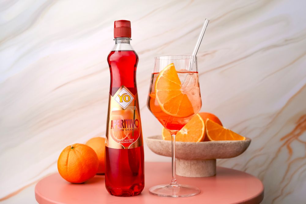 A bottle of YO Aperitivo bitter orange syrup on a pink table with a glass of the drink, ice, and orange slices, alongside whole and sliced oranges.