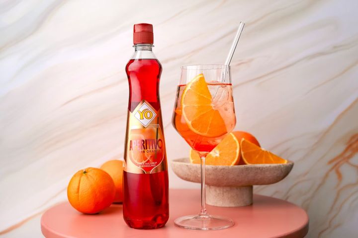 A bottle of YO Aperitivo bitter orange syrup on a pink table with a glass of the drink, ice, and orange slices, alongside whole and sliced oranges.