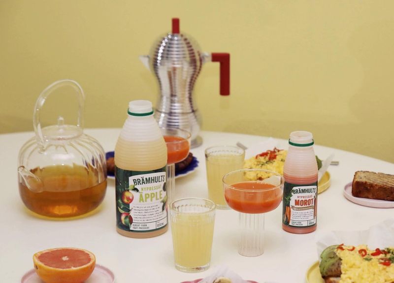 A breakfast table with apple and carrot juice bottles, a glass teapot, grapefruit halves, and light snacks including bread. A moka pot is in the background.
